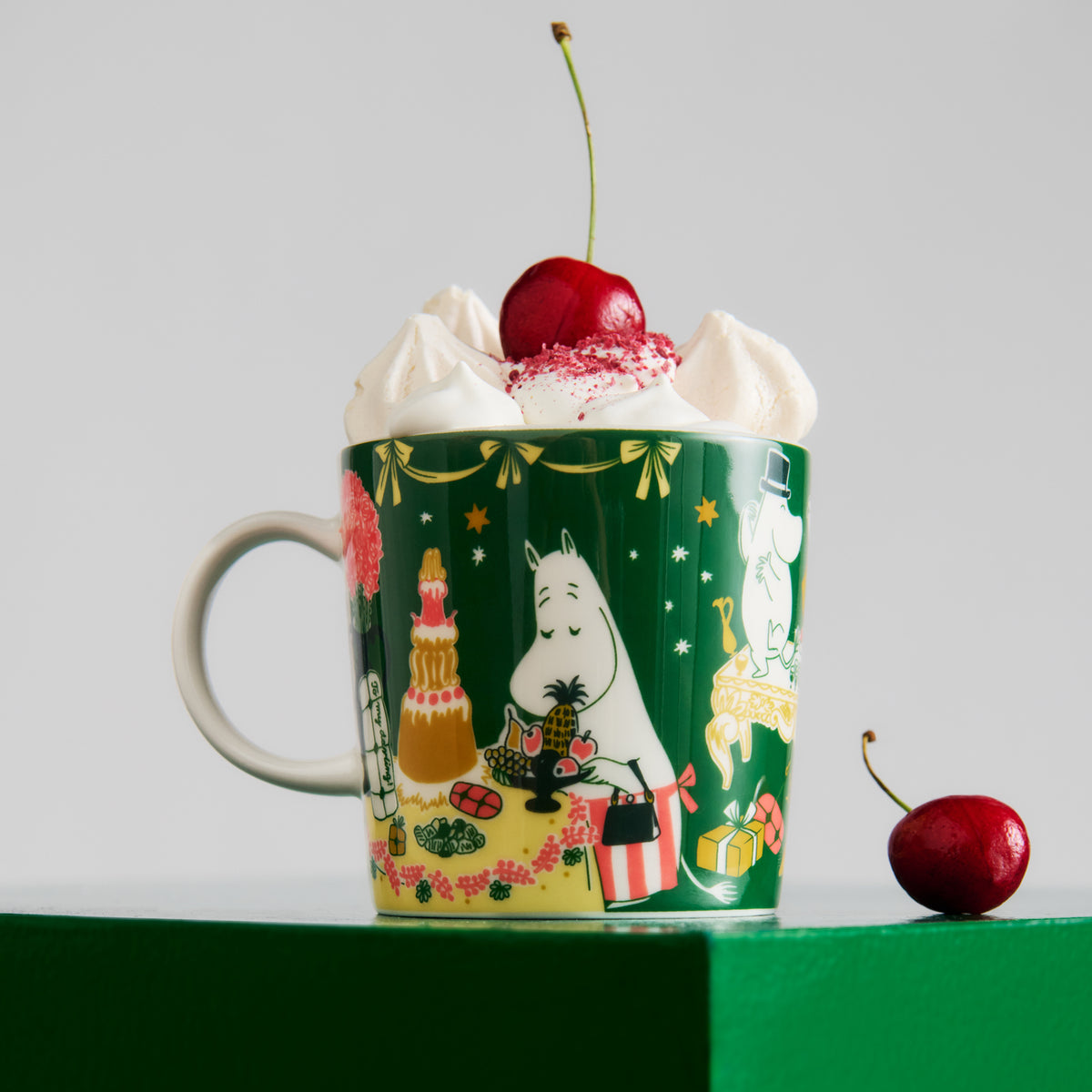 Moomin mug 0,3L Gift of Love - A green ceramic mug with Gift of Love illustrations, filled with whipped cream and topped with a cherry, set on a green pedestal against a light gray background with a second cherry on the side.