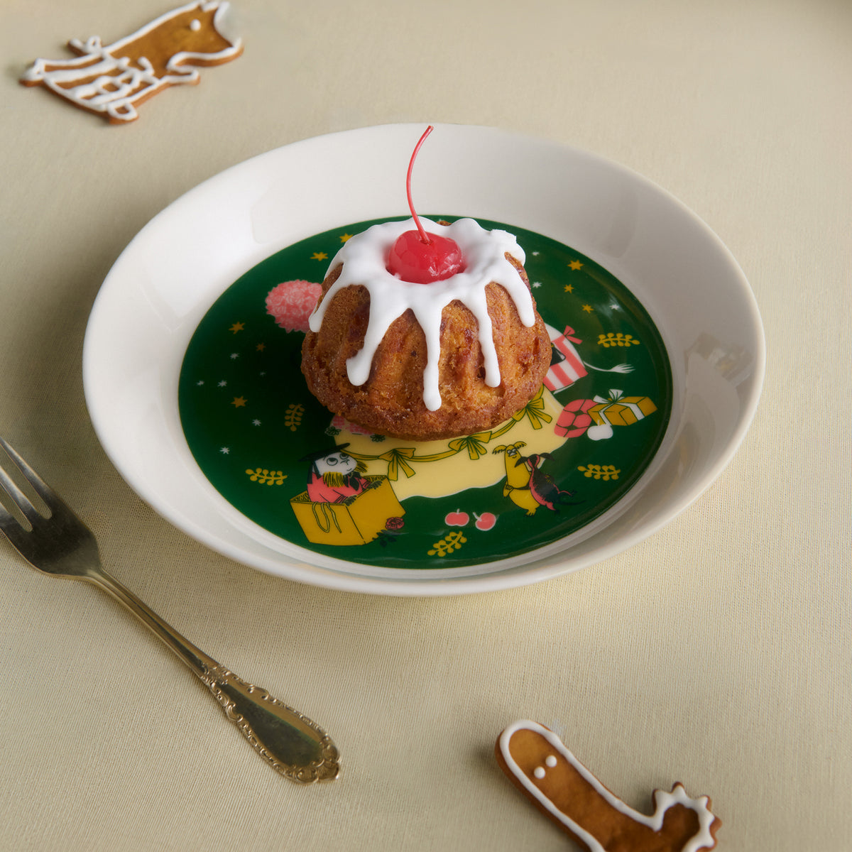 Mumin tallrik 19cm Kärleksgåva - A festive ceramic plate with a Gift of Love illustration, with a decorated pudding with white icing and a cherry on top, styled on a beige table with a fork and gingerbread cookies.