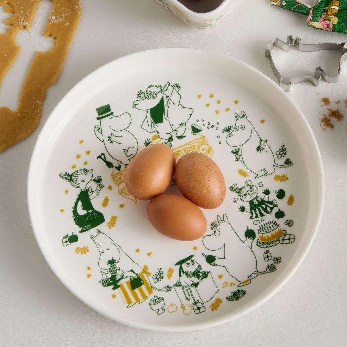 Mummi paiform 28cm Kjærlighetsgave - A white ceramic pie dish with green and gold Gift of Love illustrations, shown on a light surface with three brown eggs placed on top. Cookie cutters and cookie dough are visible around the plate.