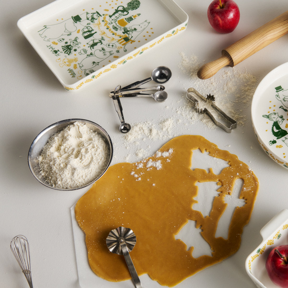 Moomin platter 24x32cm Gift of Love - A styled flat-lay of Gift of Love ceramic kitchenware on a white surface, featuring a rectangular platter with green and yellow illustrations, a matching pie dish, surrounded by baking ingredients and tools.