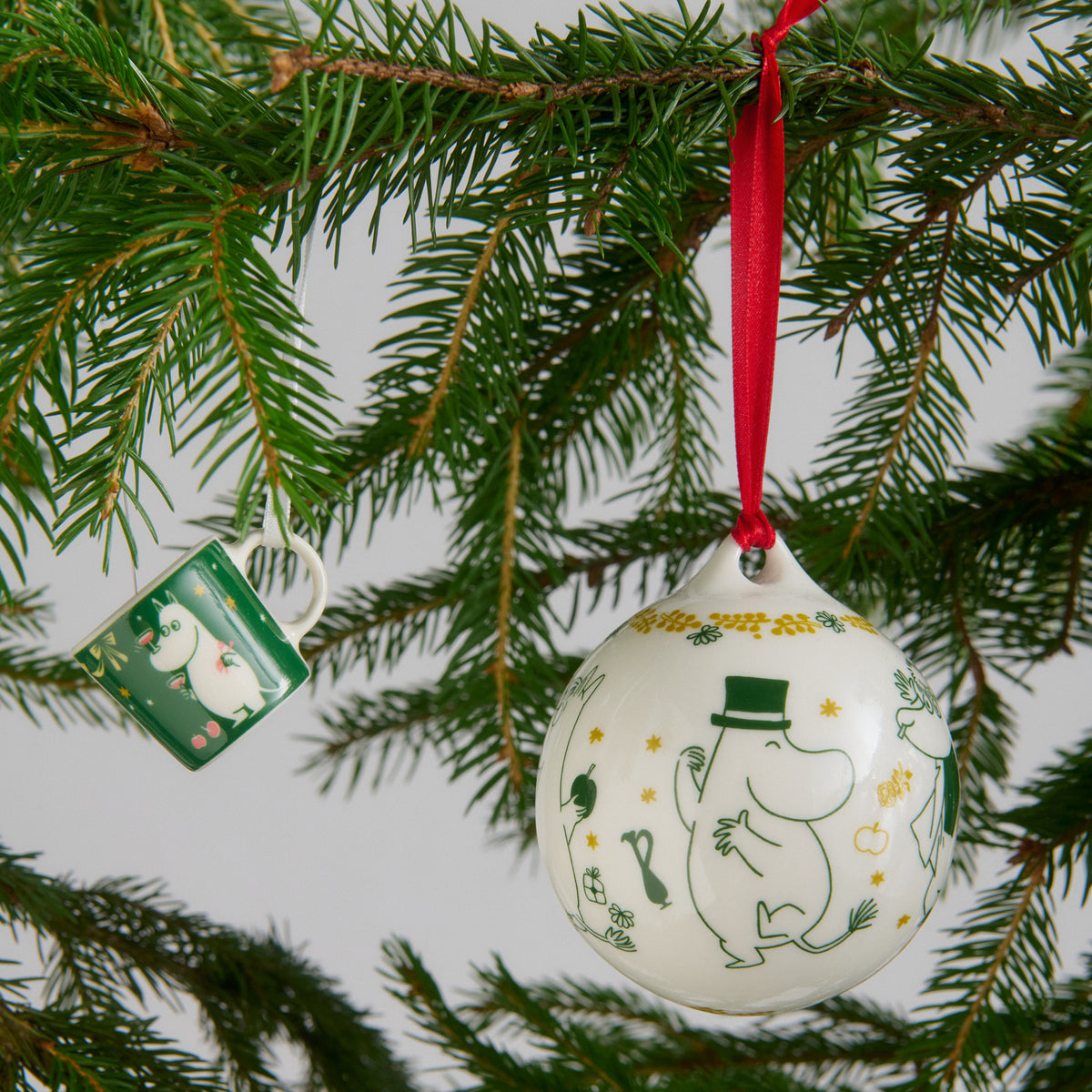 Moomin annual bauble 80mm 2026 - A white ceramic bauble featuring green and yellow Moomin character illustrations hangs from a red ribbon on a pine tree branch. A small green mini mug is visible nearby.