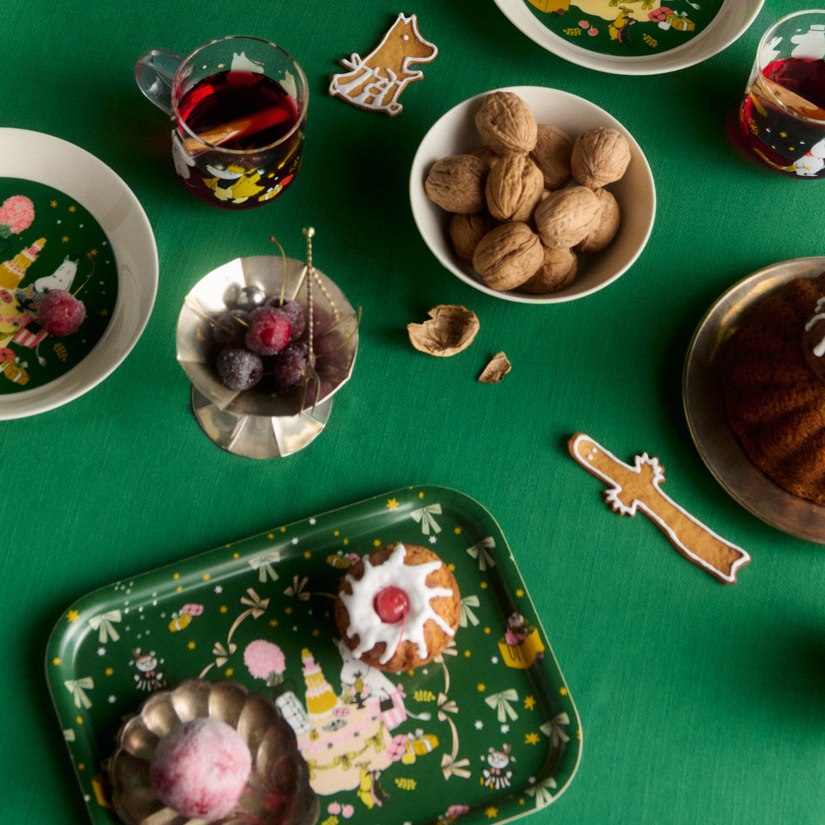 Moomin tray 27x20cm Gift of Love - A styled flat-lay of green Gift of Love tray and other products, surrounded by holiday treats like walnuts, cherries, and decorated pastries on a green tablecloth.