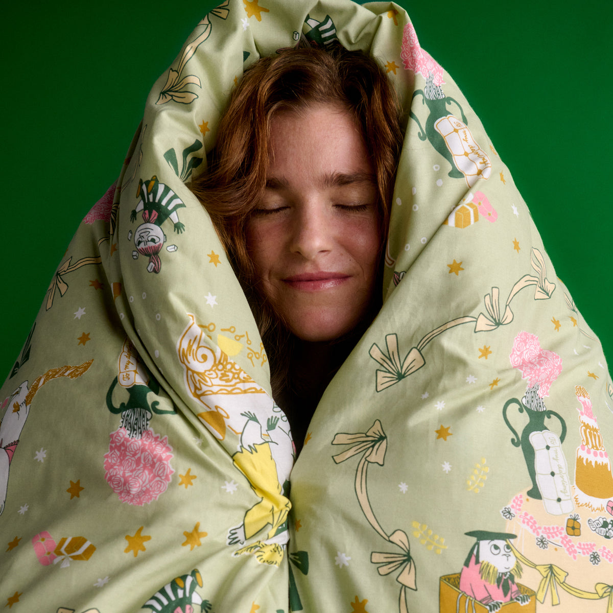 Mumin bäddset 150x210cm Kärleksgåva - A person wrapped in a light sage green patterned duvet or blanket, with their face peeking out against a solid green background. The fabric features Gift of Love illustration.