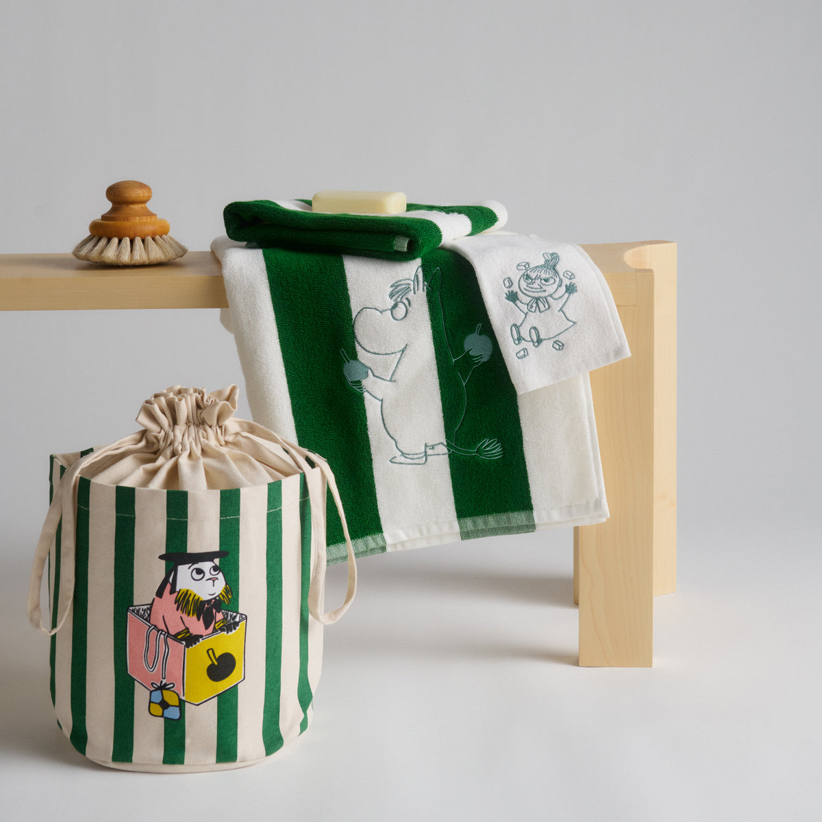 Muumi kylpypyyhe 70x140cm Lahjana Rakkaus - A set of Moomin-themed towels and a storage bag. The set includes a green and white striped towel with Moomin character illustrations, a folded green towel, and a matching green and white striped drawstring storage basket.