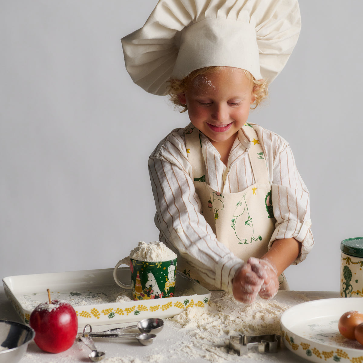 Mumin barn förkläde Kärleksgåva - A young child wearing a cream chef hat and chef apron with Gift of Love illustrations, smiling while mixing flour on a table with baking utensils, a red apple, and ceramic dishes against a light gray background.