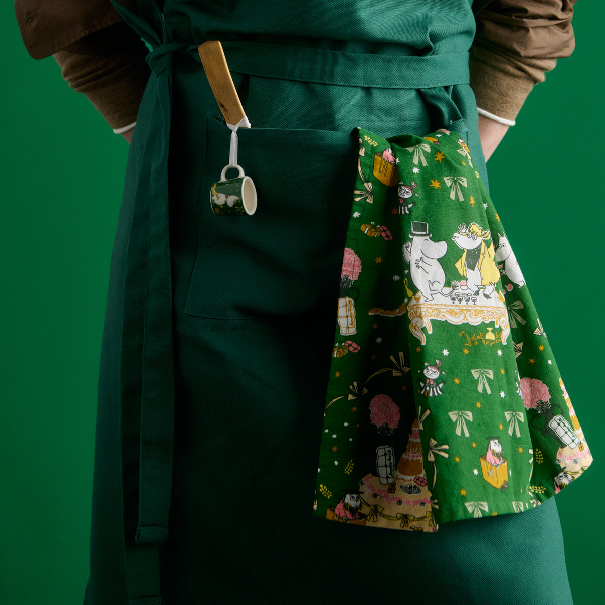 Moomin kitchen towel 50x70cm Gift of Love - A dark green apron with a front pocket, worn by a person against a solid green background. A small green ceramic mini mug with a wooden handle is clipped to the pocket, and a green patterned kitchen towel Gift of Love illustration is tucked into the side pocket.