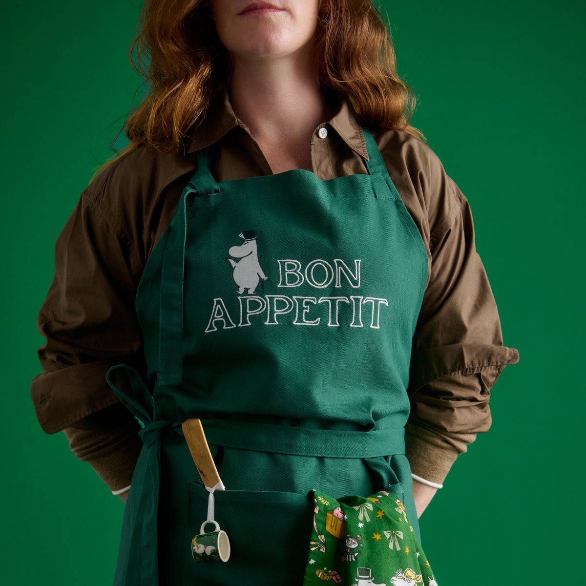 Mummi forkle Alfabet Mummipappa - A person wearing a dark green apron with 'BON APPETIT' and a Moominpappa illustration printed in white, worn over a brown button-up shirt against a solid green background.