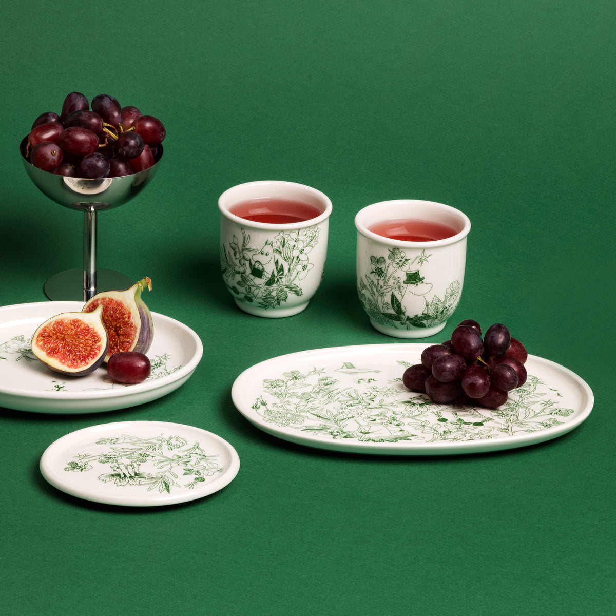 Mummi serveringsfat 17 cm Haru grønn - A set of Haru Green ceramic tableware with green botanical illustrations, arranged on a solid green background with fruit props including grapes, figs, and two cups of red drink.