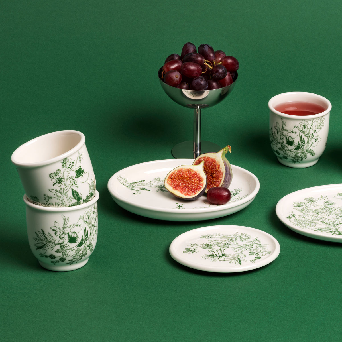 Moomin oval plate 16cm Haru green - A coordinated set of white ceramic tableware with a green botanical toile-style print, arranged on a solid green surface. The set includes stacked cups, a small cup with red beverage, plates, and a central plate holding fig halves and a grape, with a metal bowl of grapes behind it.