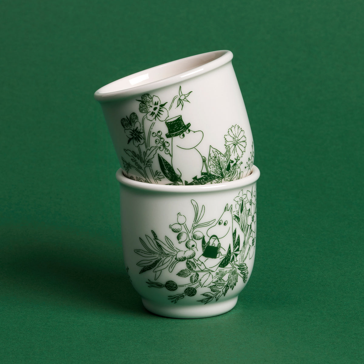 Moomin tea mug 26cl Haru green - Two stacked white tea mugs decorated with a dark green botanical and floral pattern, set against a solid dark green background.