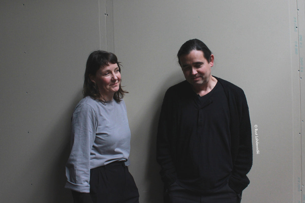 Two people standing against a plain wall.