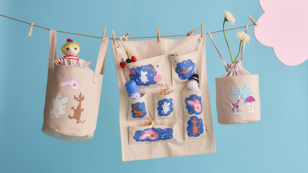 A beige fabric hanging Moomin organizer set with multiple pockets and clips, shown hanging from a string against a light blue background. The set includes a large central organizer panel with blue and multicolor animal-themed pockets, a left hanging bag with Moomin illustrations and a doll peeking out, and a right hanging bag with a unicorn and umbrella illustration.