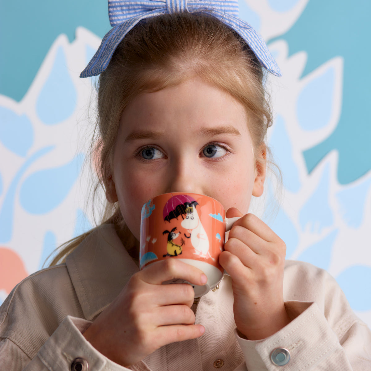 A young girl with a blue gingham bow in her hair holds a Moomin mug. She is wearing a beige coat against a light blue and white patterned background.
