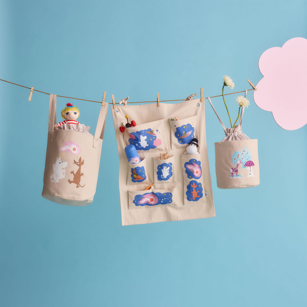 Moomin storage basket 24cm Under The Umbrella - A beige fabric storage organizer hanging from a clothesline with wooden clips, decorated with colorful patches and holding plush toys, shown against a light blue background with a pink cloud cutout.