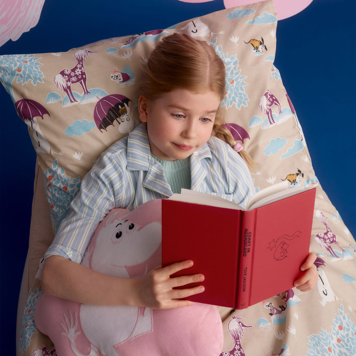 Mummi pute 37x47cm Romeventyr - A young girl with red hair lies in bed reading a red book, surrounded by matching beige bedding with Under the Umbrella illustration, and a pink plush pillow.