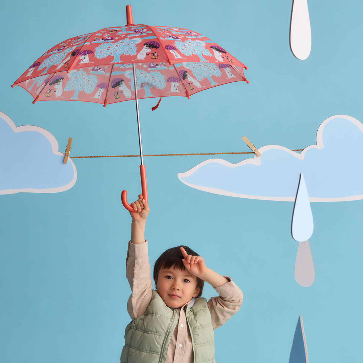 Moomin kids umbrella Under the Umbrella - A child wearing a light green puffer vest holds up a red umbrella with an Under the Umbrella print, set against a blue backdrop with paper clouds and raindrops.