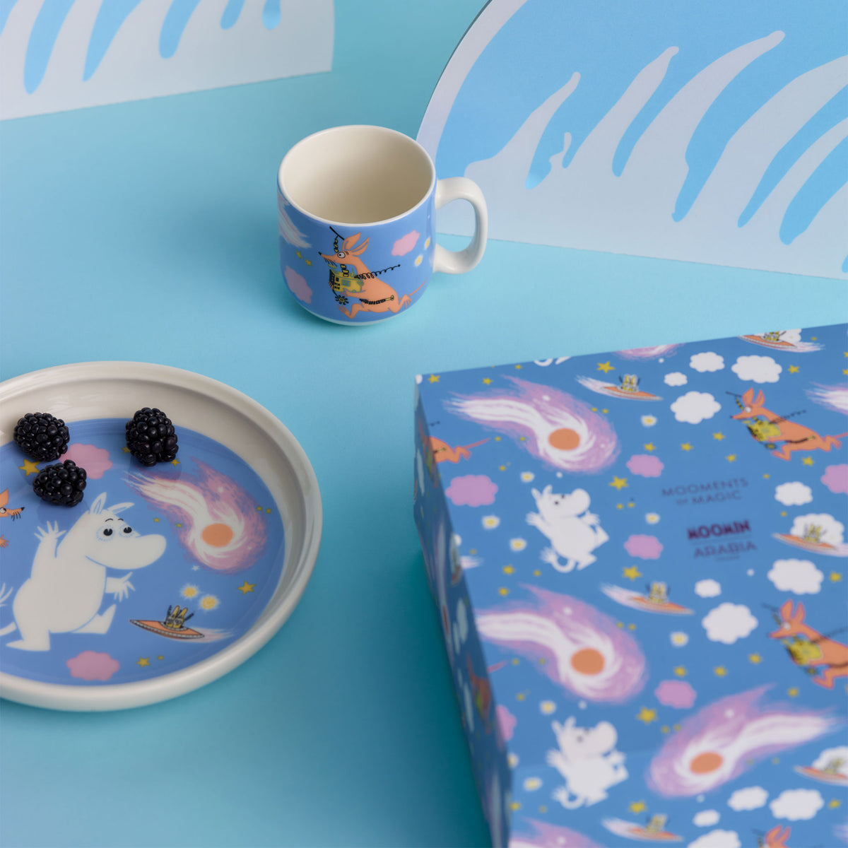 Moomin Children's set Space adventure - A Space Adventure-themed tableware set on a bright blue background, including a mug, a plate with blackberries, and a matching gift box. 
