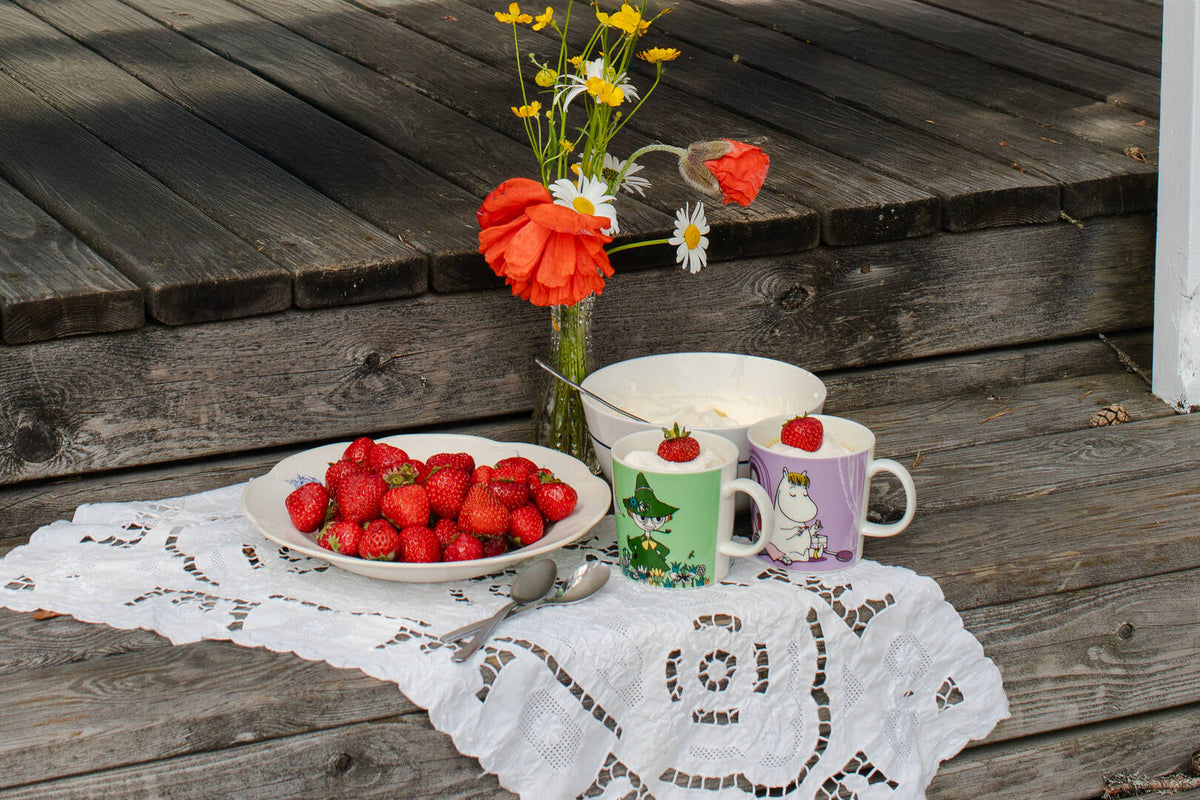 Strawberries on a plate with two Moomin mugs and a vase of flowers on a wooden surface