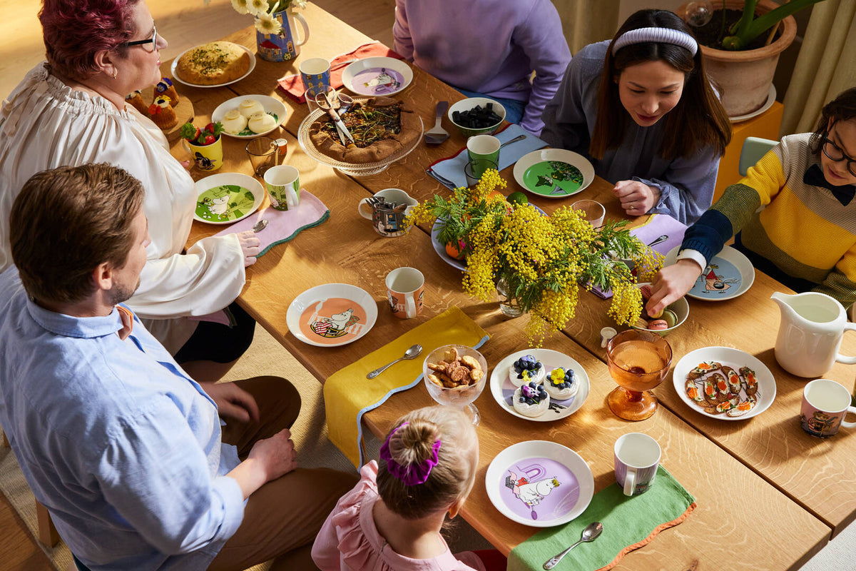 People gathered around a dining table for a meal, with Moomin classics plates and mugs on the table.