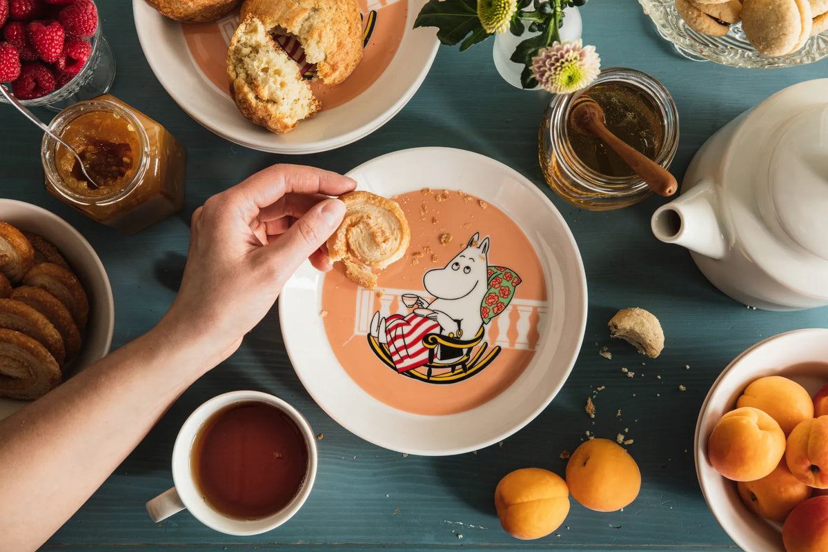 Person holding a cookie over a plate with a Moominmamma character, surrounded by tea, cookies, and fruits on a teal table.