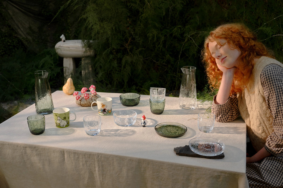 Woman with red hair sitting at a table set with Mystical forest tumblers, bowls, and vases outdoors.
