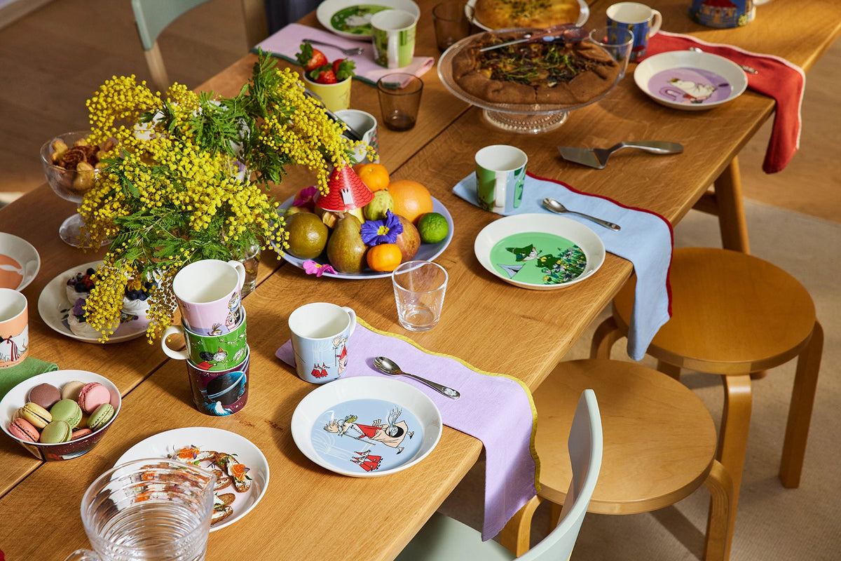 Dining table set with Moomin character design plates, cups, and food on a wooden table.