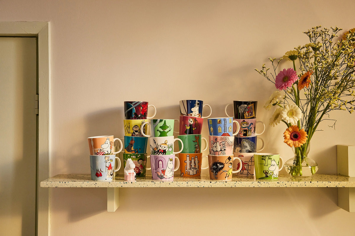 Collection of colorful Moomin classics mugs on a shelf against a beige wall with a vase of flowers.