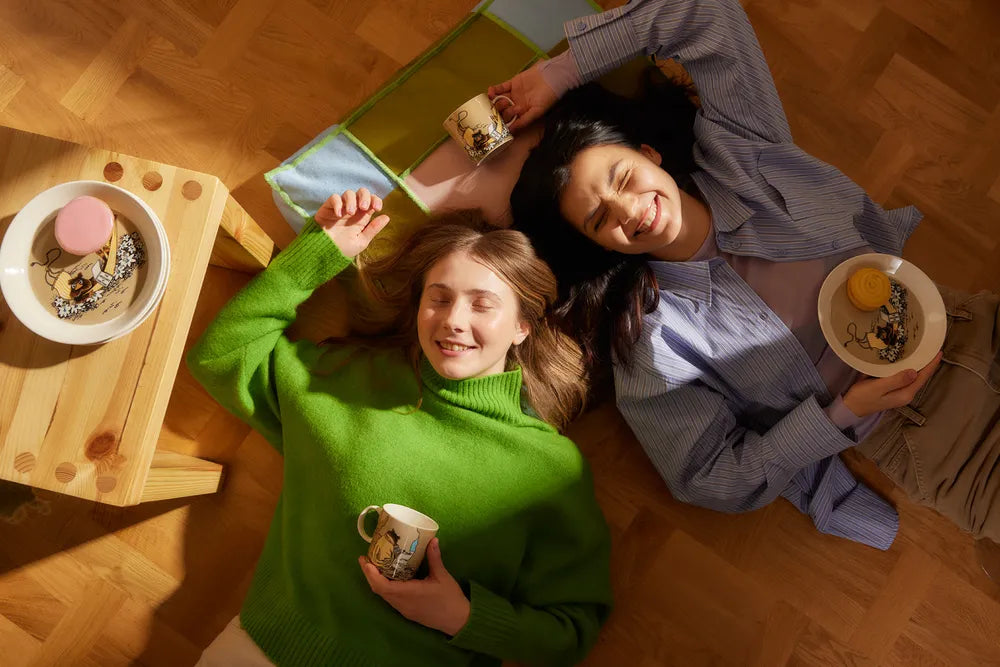 Two people lying on a wooden floor with Moomin Arabia Muskrat tableware around them